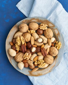 Classic blue in food. Mix of nuts on a plate - walnut, almonds, pecans, macadamia and knife for opening shell. Healthy vegan food. Clean eating, balanced diet. Top view, vertical