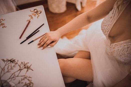 The girl sits with her legs crossed at a white cosmetic table. The bride in a white wedding dress with an open neckline. Various professional cosmetics hairbrush female accessories laid out on the tab