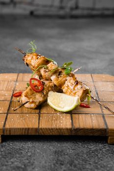 Meat on wooden skewers, garnished with cilantro, lime, sesame, chili on square board on gray background