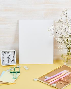 Mockup with blank white frame on yellow table against wooden wall, alarm, flower in vaze, vertical.