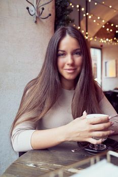 Beautiful pensive happy girl sitting in cafe in Christmas holidays, smiling and dreaming. Brunette woman with long hair drinks cappuccino coffee, latte and looks to camera, vertical