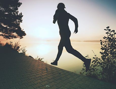 Freeze action shot of tall running sportsman wearing trendy running shoes and sports clothes exercising morning at shore. Pavement track around lake.