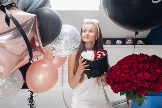 Young girl laughing and enjoying beautiful presents at home. Happy woman sitting neat big balloons and big bouque of red roses, keeping heart of flowers and posing. Concept of happiness and love.