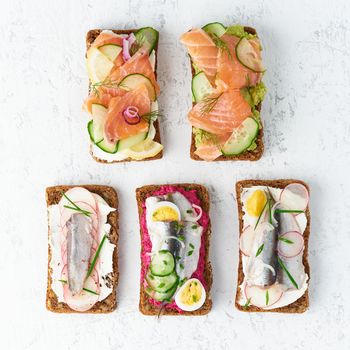 Savory fish smorrebrod, set of five traditional Danish sandwiches. Black rye bread with anchovy, beetroot, radish, eggs, salmon, cream cheese, cucumber, avocado on a grey white stone table, top view