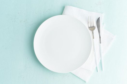 Clean empty white plate, fork and knife on turquoise green blue stone table, copy space, mock up, top view. Concept for menu with utensil