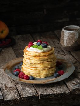 Pancake with vanilla cream, blueberries and raspberries. Side view, vertical. Dark moody old rustic wooden background.