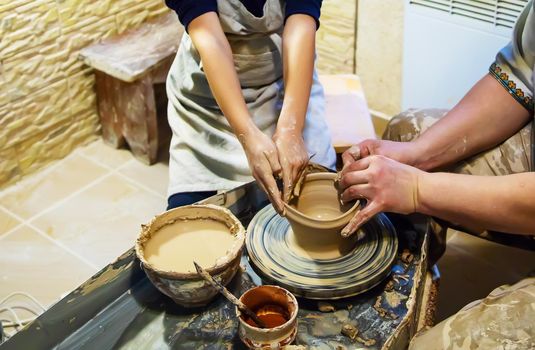 the Hands of a master and a student make a pitcher on a Potters wheel of yellow clay. Selective focus on hands