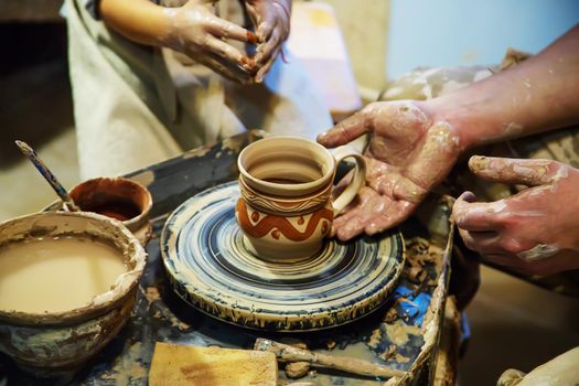 the Hands of a master and a student make a pitcher on a Potters wheel of yellow clay. Selective focus on hands