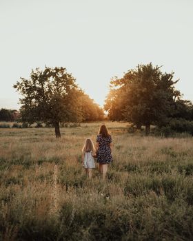 European family mom and daughter walk holding hands Along the tall green grass towards the sunset in summer. meadow and tall trees through which the rays of the sun make their way