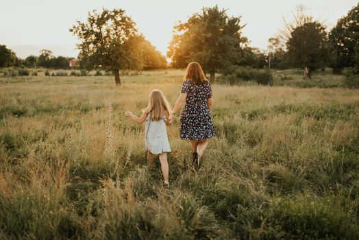 European family mom and daughter walk holding hands Along the tall green grass towards the sunset in summer. meadow and tall trees through which the rays of the sun make their way