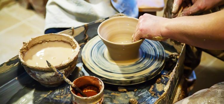 the Hands of a master and a student make a pitcher on a Potters wheel of yellow clay. Selective focus on hands