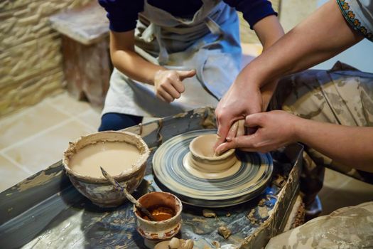 the Hands of a master and a student make a pitcher on a Potters wheel of yellow clay. Selective focus on hands