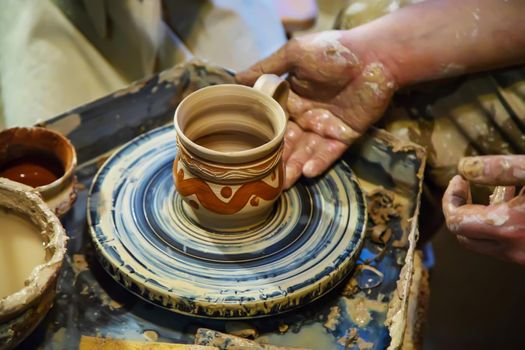 the Hands of a master and a student make a pitcher on a Potters wheel of yellow clay. Selective focus on hands