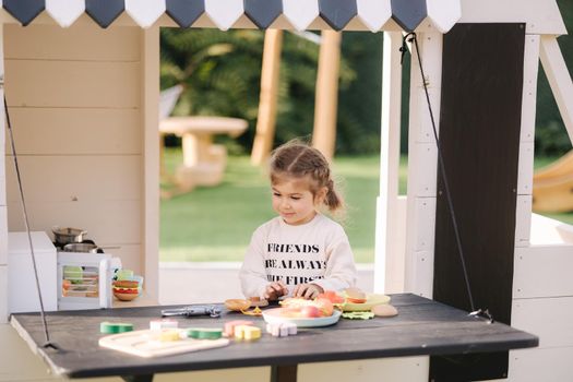 Happy little girl playing on toy kitchen on wheels. Cute girl make a burger on toy kitchen outdoors. Adorable kid play on fresh air.