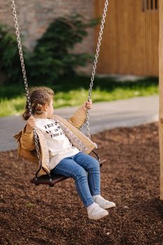 Happy little girl swing on a swing in the specially designated place in the park. Softner from sawdust underfoot.