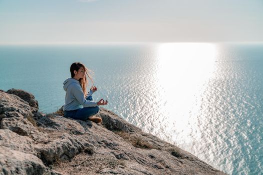 Woman tourist enjoying the sunset over the sea mountain landscape. Sits outdoors on a rock above the sea. She is wearing jeans and a blue hoodie. Healthy lifestyle, harmony and meditation.