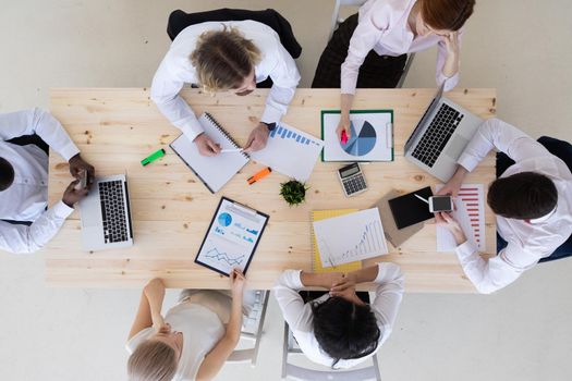 Top view of business people in formal wear sitting in boardroom at desk and analyzing data. On desk are laptops, graphs, tablets and paperwork.