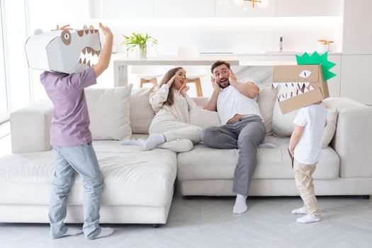 Happy family of parents and two children playing dinosaurs at home, children wearing handmade costume mask of cardboard