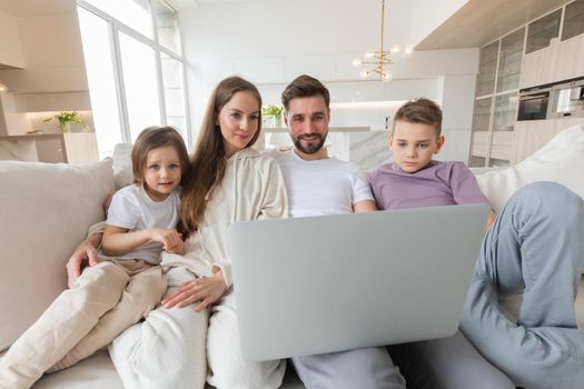 Happy young family with little kids sit on sofa in living room using modern laptop together, smiling parents rest on couch enjoy weekend with small children laugh watch video on computer at home