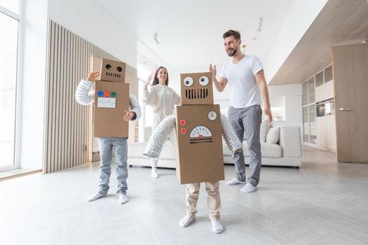 Happy family of parents and two children playing dancing like robots at home, children wearing handmade moving box costume of cardboard