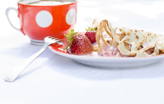 Pancakes with strawberries and sour cream on the white tablecloth. Next to a cup of coffee. All lit bright sun. On the tablecloth visible shadows from tree leaves.