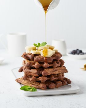 Chocolate banana waffles with maple syrup on white table, side view, vertical. Sweet brunch, maple syrup flow in milk jug, a creamer.