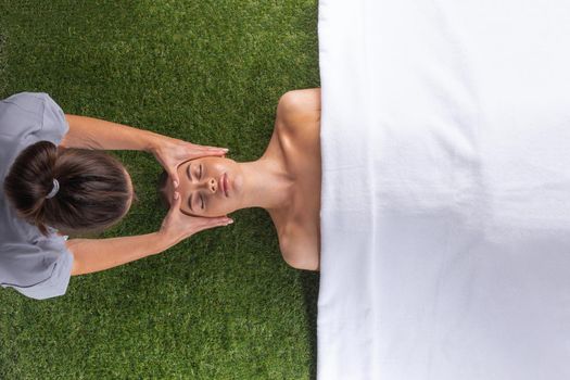 Releasing stress. Top view of beautiful young woman lying on back while massage therapist massaging her face over green grass background