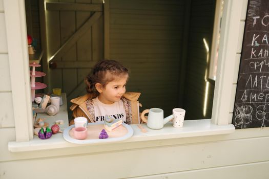 Little girl sits inside of a toy cafe. Happy kid playing with toys outdoor. Little toy city for children.