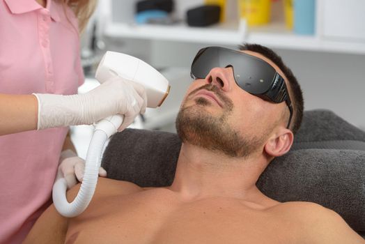 Laser hair removal on mans face. man in a goggles. therapist in a pink t-shirt