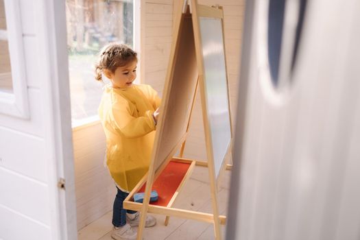 Young artist paints on wooden boards with chalk. Cute girl in yeallow raincoat.