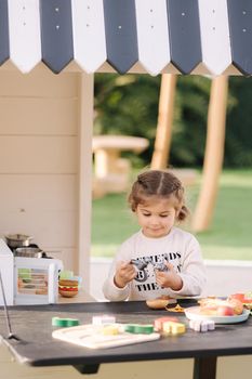 Happy little girl playing on toy kitchen on wheels. Cute girl make a burger on toy kitchen outdoors. Adorable kid play on fresh air.