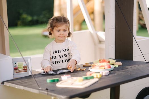 Happy little girl playing on toy kitchen on wheels. Cute girl make a burger on toy kitchen outdoors. Adorable kid play on fresh air.