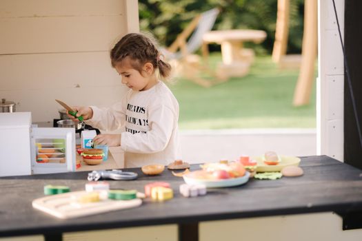 Happy little girl playing on toy kitchen on wheels. Cute girl make a burger on toy kitchen outdoors. Adorable kid play on fresh air.