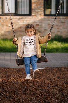 Happy little girl swing on a swing in the specially designated place in the park. Softner from sawdust underfoot.