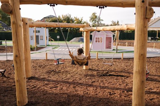 Happy little girl swing on a swing in the specially designated place in the park. Softner from sawdust underfoot.