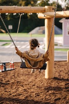 Happy little girl swing on a swing in the specially designated place in the park. Softner from sawdust underfoot.