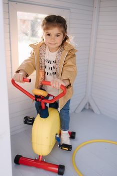 Happy little girl have sports in a toy gym. Liliput fitness center. Cute girl sits on children exercise bike and smile.