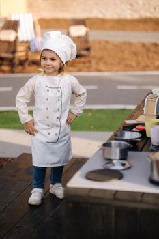 Adorable little girl in chef's coat and cap cooks at the children's toy kitchen. Playing on little kids city.