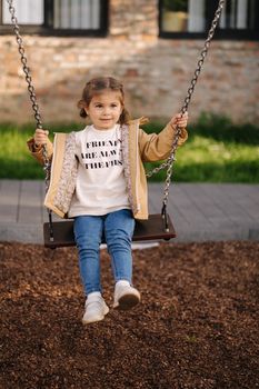 Happy little girl swing on a swing in the specially designated place in the park. Softner from sawdust underfoot.