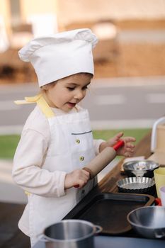 Adorable little girl in chef's coat and cap cooks at the children's toy kitchen. Playing on little kids city.