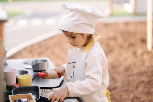 Adorable little girl in chef's coat and cap cooks at the children's toy kitchen. Playing on little kids city.