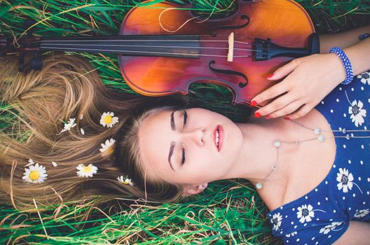 young slim fair-skinned girl blonde beautiful lies in tall grass. Girl in a blue short tight dress with a print of daisies. A bouquet and lonely daisies scattered around.A woman hugs violin. Top view