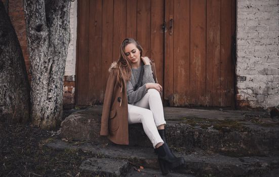 beautiful young blonde model girl. White pants. gray knitted sweater. boots. wooden pendant on the neck in the form of a horse. in a brown coat posing. On the Sunset. Portrait. sitting on the steps