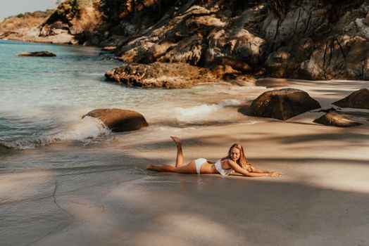 young blond European woman in white bikini swimsuit on beach lies on sand beach sea. background of large stone rocks and palm trees