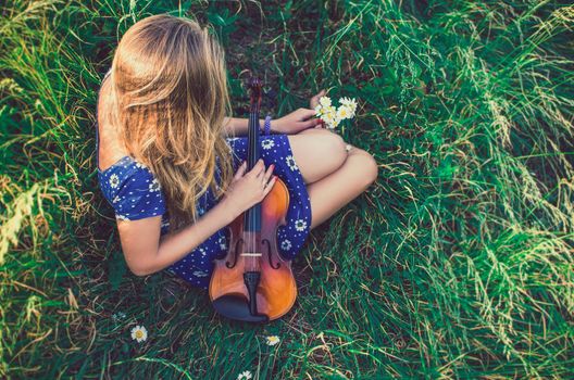 Young slim fair-skinned girl blonde beautiful sit in tall grass. Girl in a blue short tight dress with a print of daisies. A bouquet daisies. A woman hugs violin. Top view