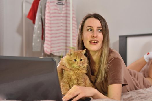 Young caucasian brunette businesswoman working at home using laptop while caressing pet red cat