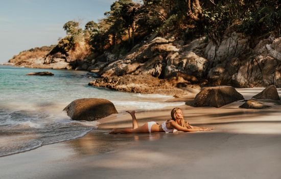 young blond European woman in white bikini swimsuit on beach lies on sand beach sea. background of large stone rocks and palm trees