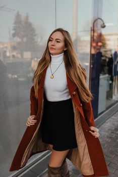 young blonde woman in autumn clothes walks on street background shop windows. Street style Fashion 2021 Winter Spring Terracotta Coat Woolen Cashmere Suede Over Knee Boots White Golf