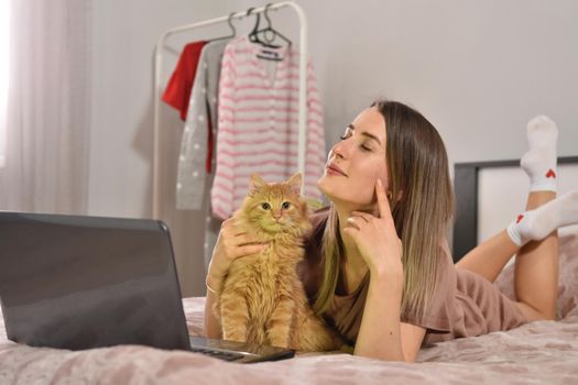 Young caucasian brunette businesswoman working at home using laptop while caressing pet red cat