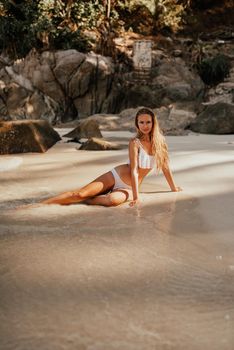 young blond European woman in white bikini swimsuit on beach sitting on sand beach, sea on background of large stone rocks and palm trees